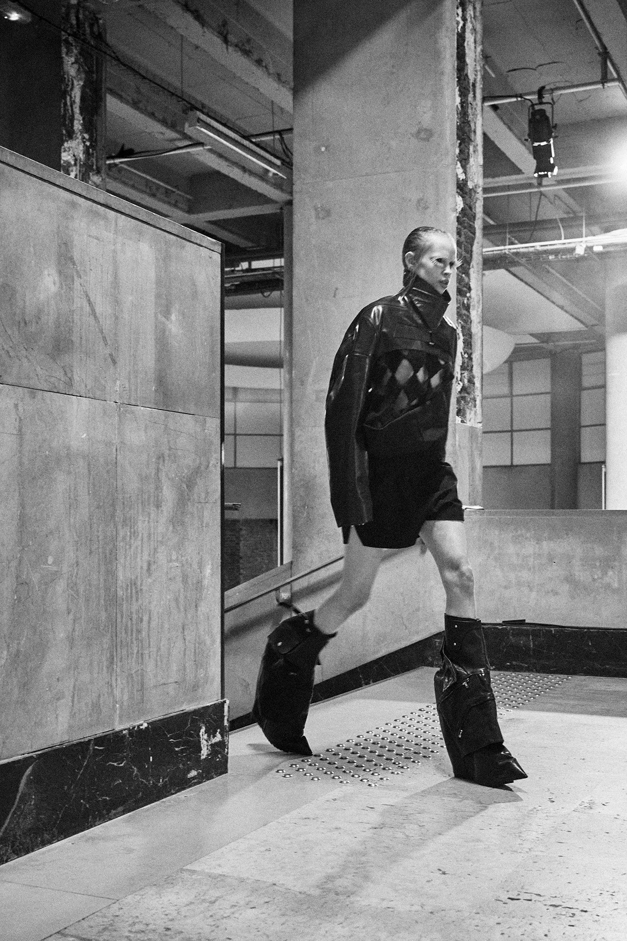 Silhouette en marche dans un hall de béton : veste de cuir courte à losanges, short, bottes plates-formes très hautes.